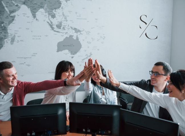 Equipo de trabajo de SOPHIA CONSULTORES celebrando con un 'high five' frente a monitores de computadora, con un mapa del mundo en la pared de fondo y el logo de la empresa visible en la esquina superior derecha.
